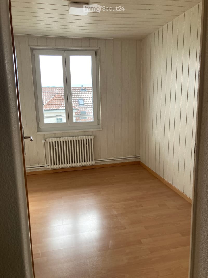 Empty room with wooden flooring, light wooden panel walls, a window with white frame, and a radiator below it.