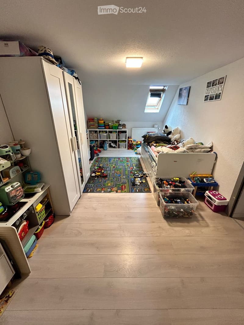 A well-lit children's room with wooden flooring, a large wardrobe, shelves filled with toys, a toy car track, and a bed with stuffed animals.