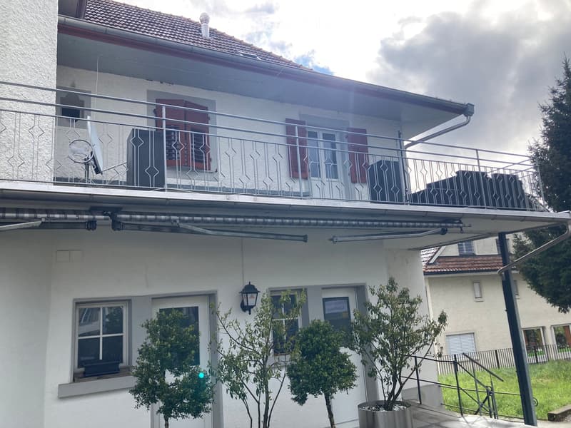 Two-story house with white exterior, red shutters, balcony with metal railing, small trees in pots, and a covered entrance area.