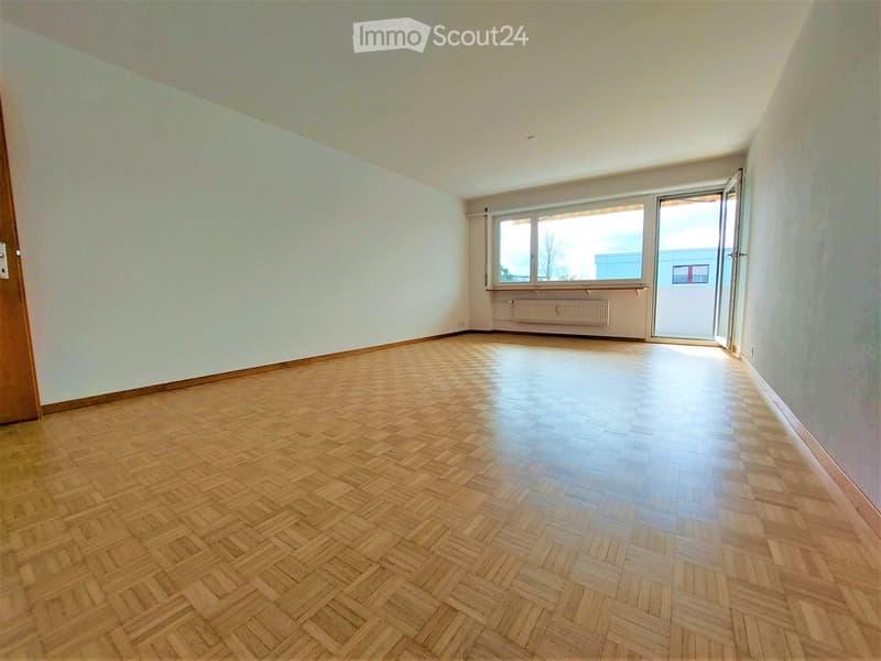 Empty room with wooden herringbone flooring, white walls, large window, and open door leading outside.