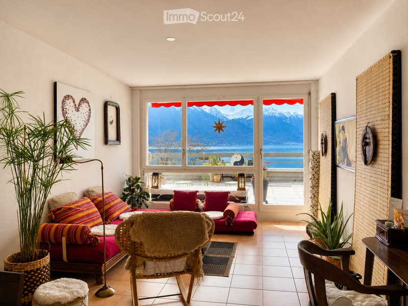 Bright living room with large windows offering a scenic mountain view, featuring a red sofa with colorful pillows, a wicker chair with a fur throw, a small table, and potted plants.