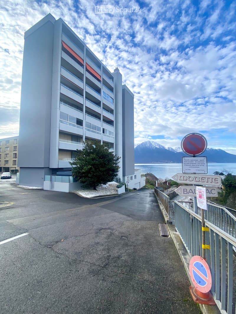 A modern multi-story apartment building with balconies and large windows, situated near a waterfront with mountains in the background.