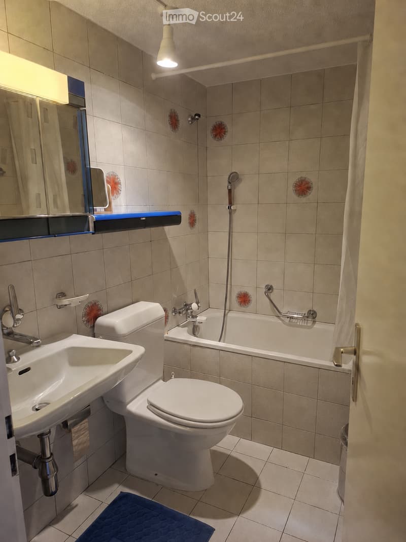 Modern bathroom with beige tiled walls and floor, featuring a sink, toilet, bathtub, mirror, and a handheld showerhead. A blue bath mat is placed on the floor.