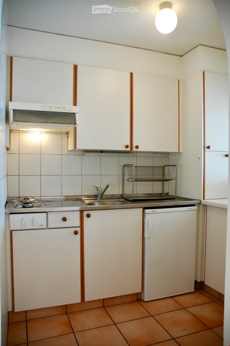 Compact kitchen with white cabinets, stainless steel sink, dishwasher, and stove. Features tiled floor and walls, and a dish rack on the counter.