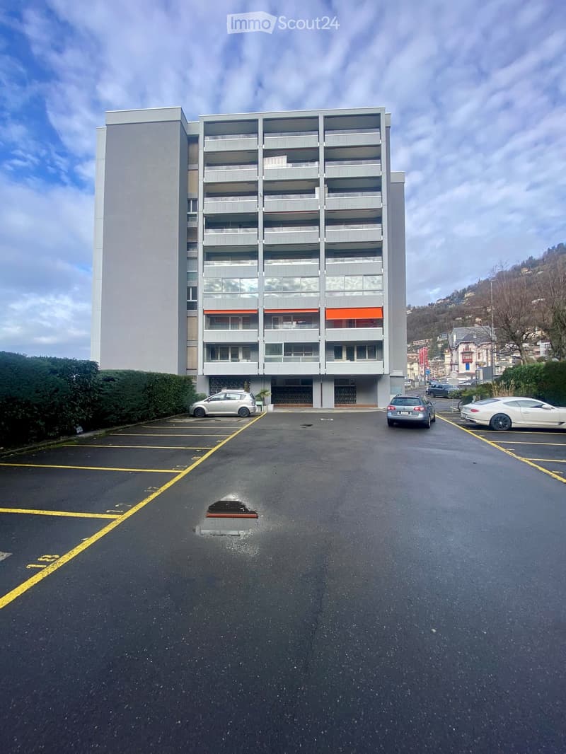 Modern multi-story apartment building with balconies and large windows, surrounded by a parking lot with several cars.