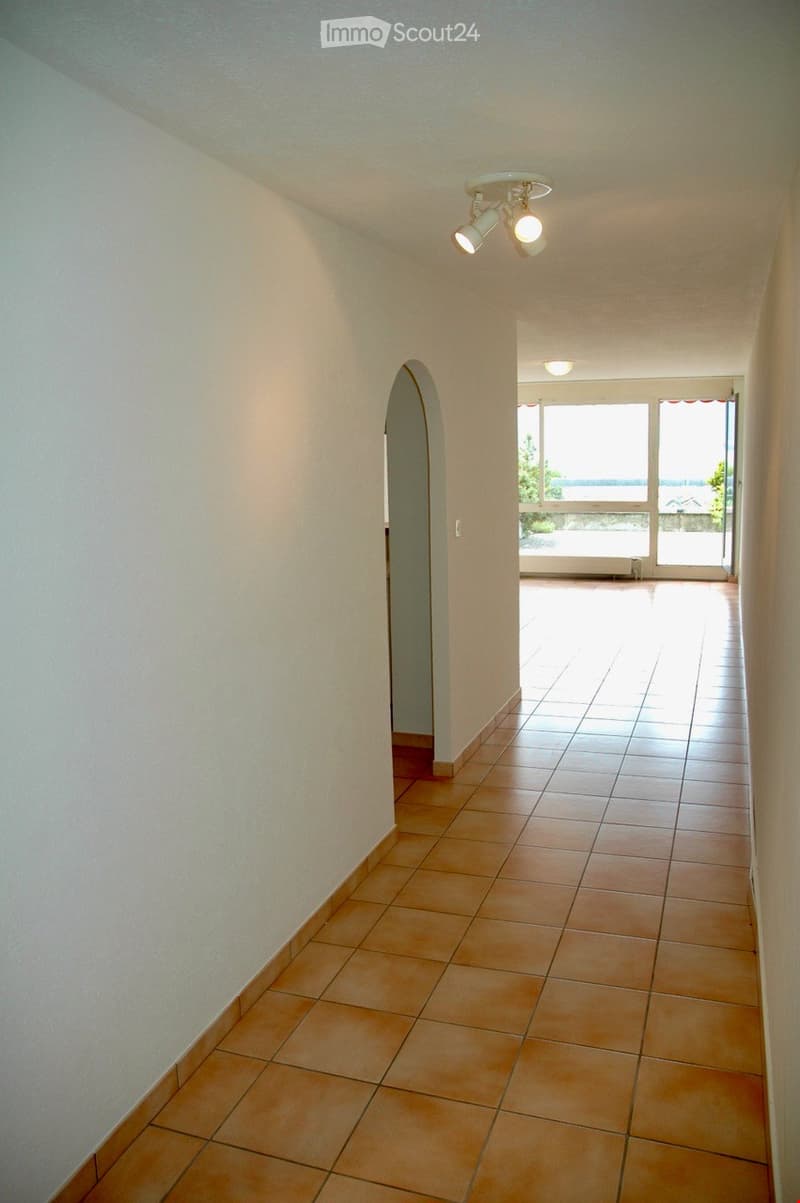 Empty hallway with beige tiled floor, white walls, and recessed lighting. An arched doorway leads to a bright room with large windows and a view of the sea.