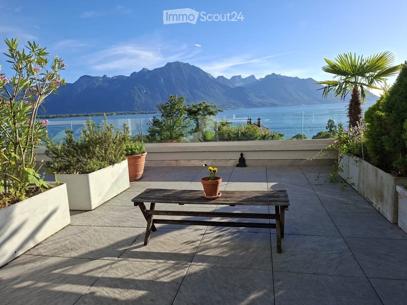 Balcony with a wooden table, potted plants, and a scenic mountain and lake view.