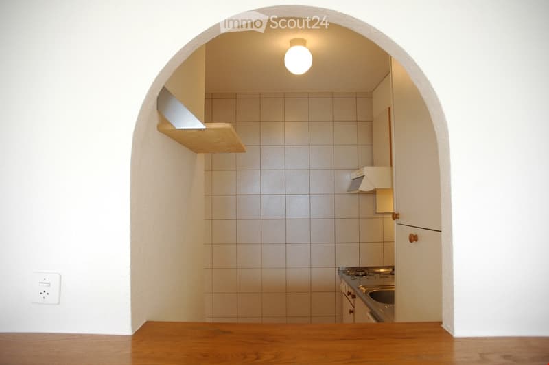 Compact kitchen with tiled walls, a stove, sink, and wooden shelf above the counter.