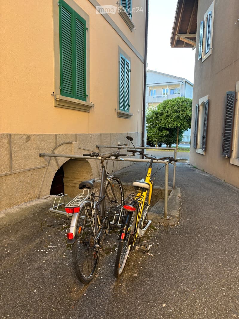 Two bicycles parked on a rack in a narrow alleyway between two buildings with green shutters and beige walls.