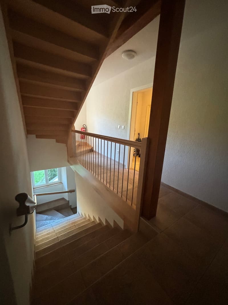 Spacious staircase with wooden steps and a metal railing, leading to an open area with a visible room beyond. Natural light streams in from a window on the left side.