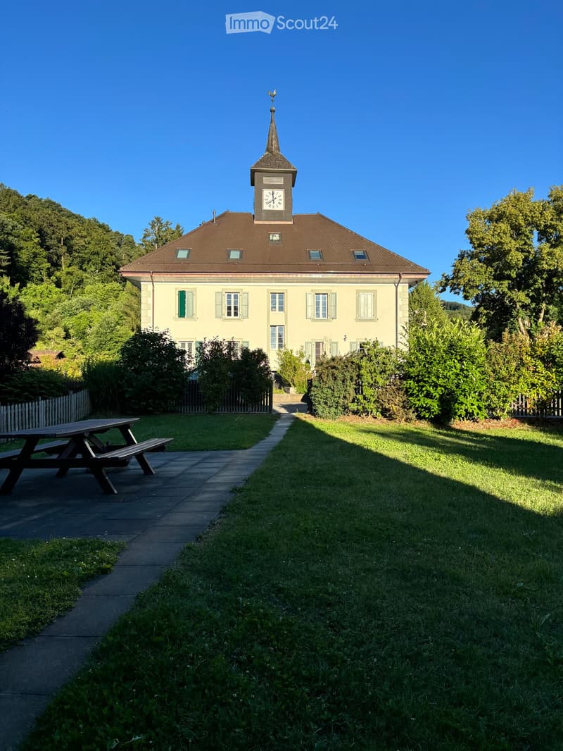 A two-story building with a clock tower, surrounded by a well-maintained lawn and trees. A picnic table is visible in the foreground.
