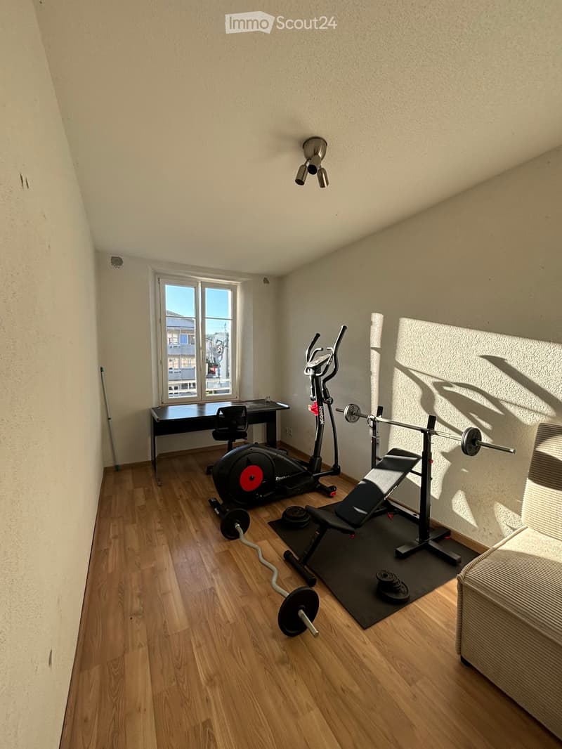 A well-lit room with wooden flooring, featuring an elliptical trainer, a bench press, dumbbells, a barbell, a chair, and a table near a large window.