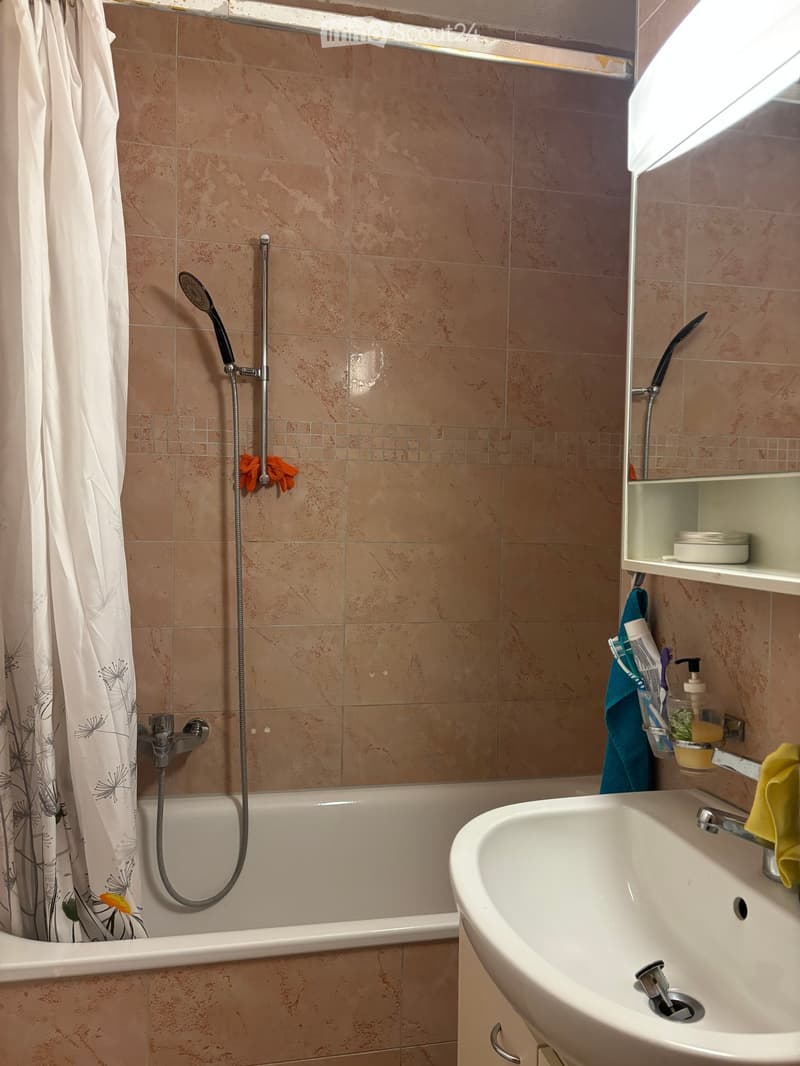 Bathroom with beige tiled walls, a bathtub with a handheld showerhead, a sink with a faucet, a shelf with toiletries, and a shower curtain.