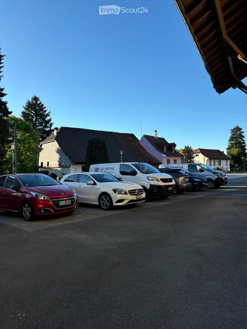 Residential area with several parked cars, including a red Peugeot, a white Mercedes, and a white van labeled "LA BROYE". Houses with pitched roofs and chimneys are visible in the background.