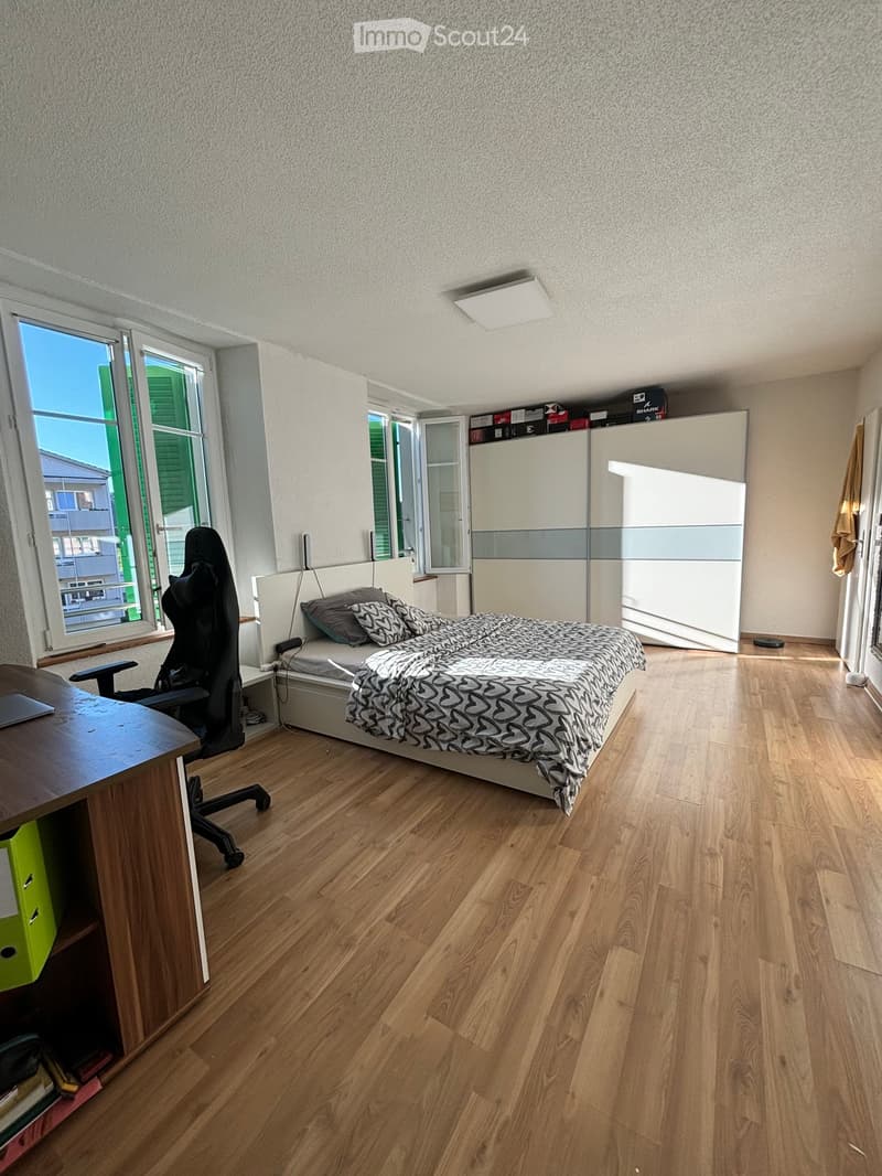 A modern bedroom with wooden flooring, a bed with patterned bedding, a desk with a chair, and a large wardrobe. The room has large windows with green shutters, allowing natural light to fill the space.