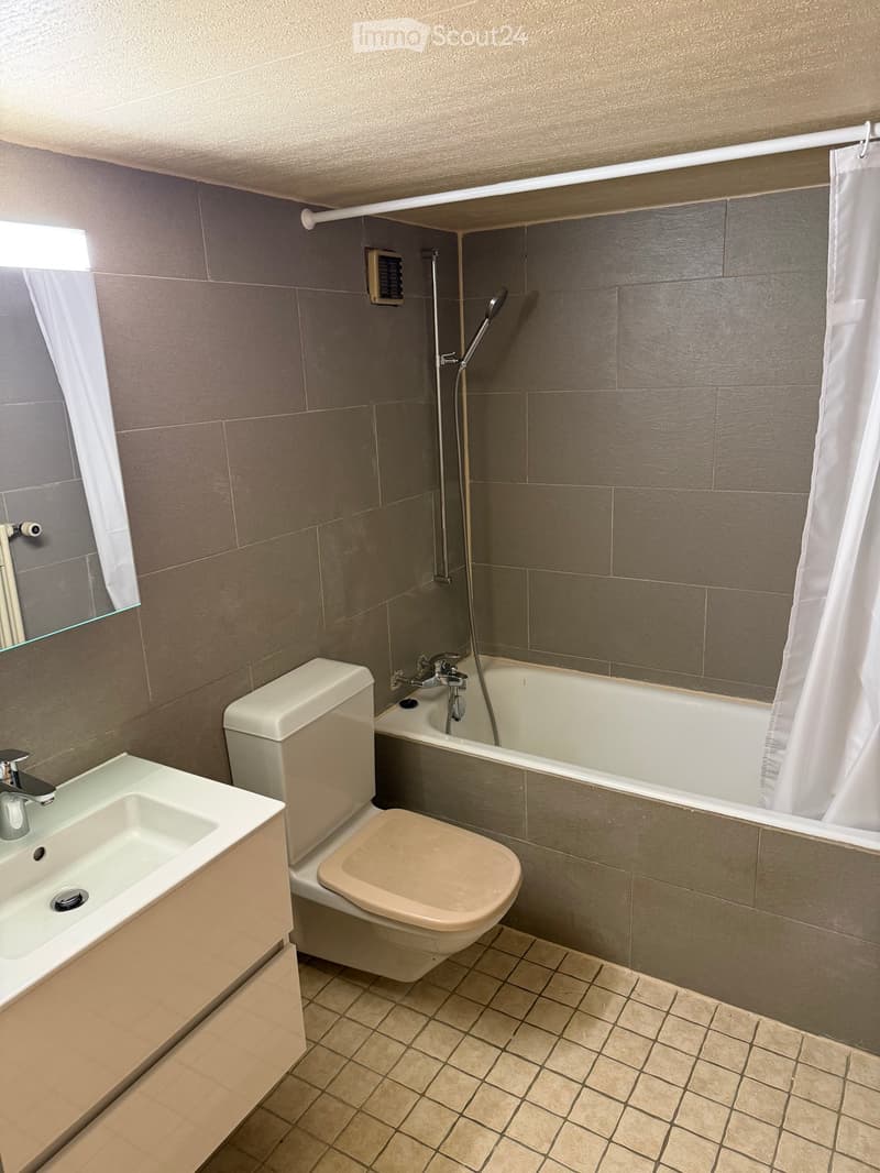 Modern bathroom with a white sink, toilet, bathtub, and showerhead. Grey tiled walls and beige tiled floor.