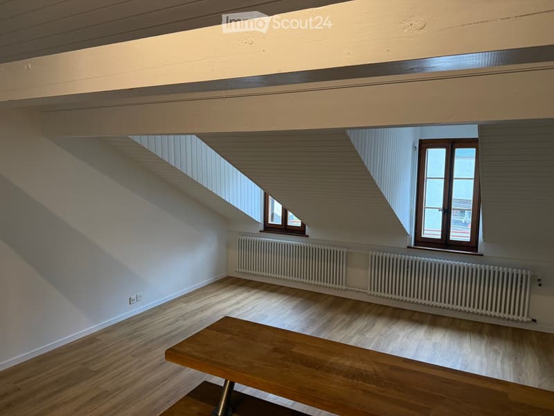 Empty attic room with wooden flooring, white walls, exposed wooden beams, and two windows with wooden frames. Radiators are present along the walls.