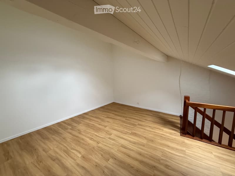 Empty room with wooden flooring, white walls, and a wooden staircase railing visible in the corner. The ceiling has exposed beams and a skylight letting in natural light.