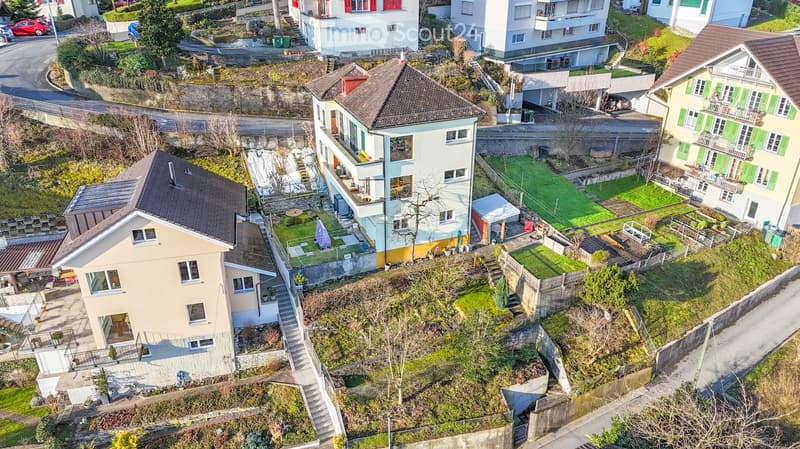 Aerial view of a residential area with multiple houses, gardens, and a winding road. Houses have varied architectural styles, with some featuring balconies and terraces. Green spaces and small gardens are visible between the buildings.