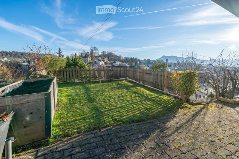 A sunny rooftop garden with a wooden fence, grassy area, and a view of the city and mountains.