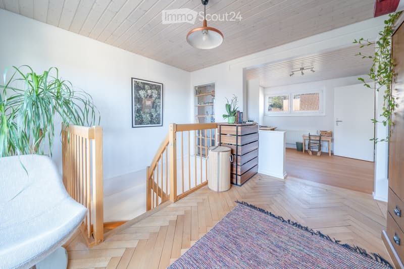 Bright living room with wooden flooring, white walls, and a wooden staircase railing. Features include a sofa, a colorful rug, a picture frame on the wall, a shelf with books and plants, a small table with a chair, and a potted plant near the window.