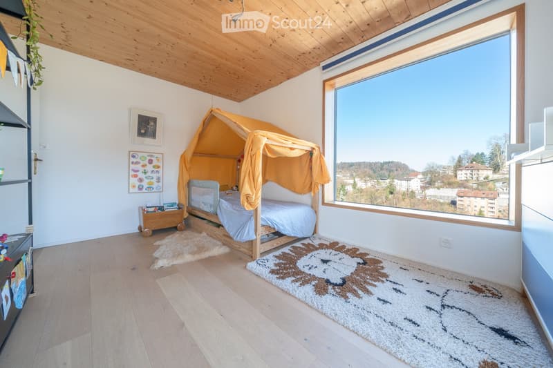 Bright bedroom with wooden floor, large window offering a scenic view, a bed with a yellow canopy, a rug, and a small cabinet with toys.