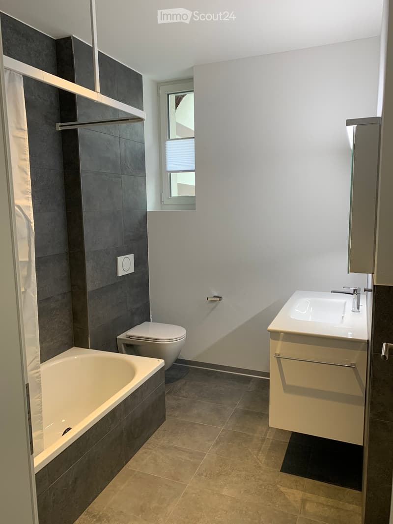 Modern bathroom with a bathtub, toilet, sink, and a window with blinds. The walls are tiled in dark gray, and the floor is tiled in beige.