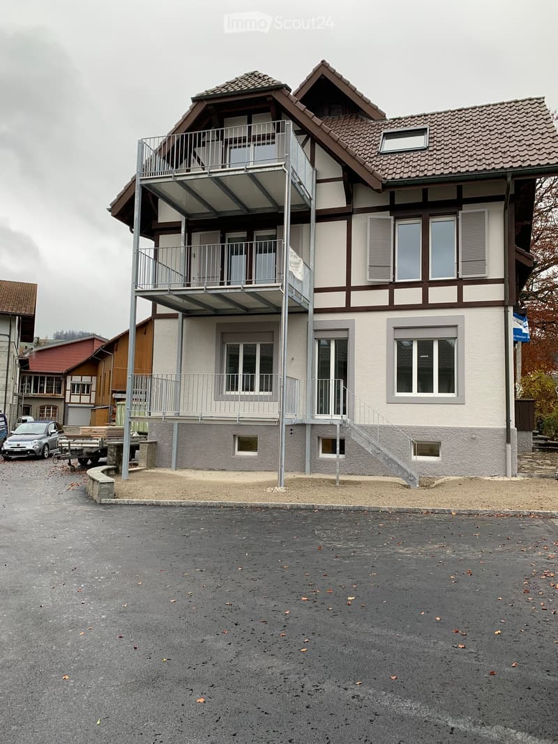 Two-story house with a modern balcony addition, traditional half-timbered upper level, and multiple windows. The ground floor has a raised entrance and a small parking area.