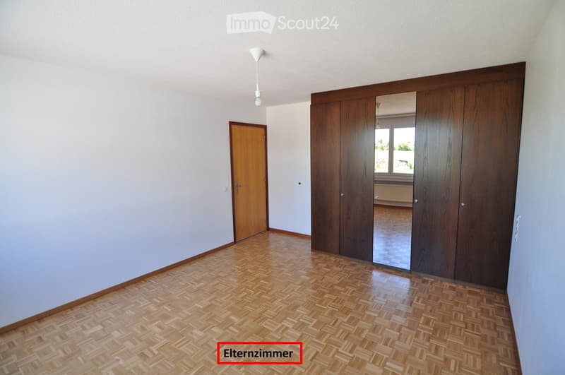 Empty room with wooden flooring, light-colored walls, and a large wooden closet with mirrored doors. A single door leads to another room.