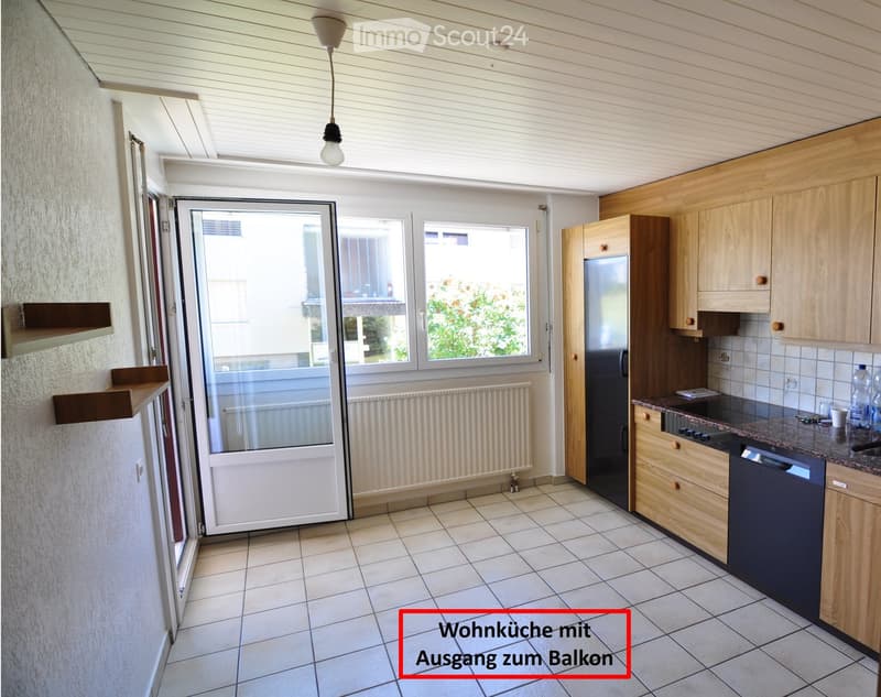 Modern kitchen with light wooden cabinets, a refrigerator, dishwasher, and a tiled floor. Features a large window and a door leading to a balcony.