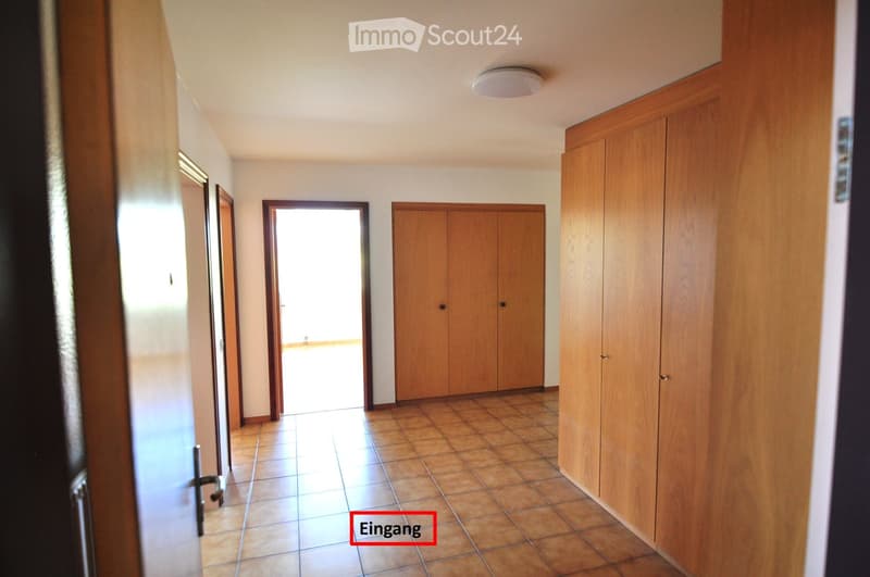Empty hallway with tiled floor, wooden cabinets, and a ceiling light. The hallway leads to an open area with natural light.