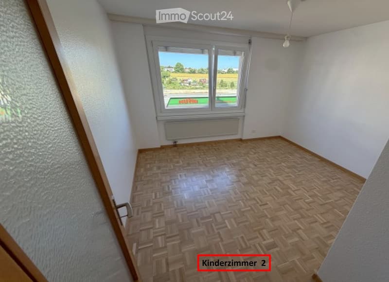 Empty room with wooden parquet flooring, white walls, large window with view, radiator, and ceiling light fixture.