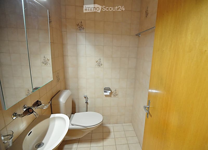 A small bathroom with beige tiled walls and floor, featuring a toilet, sink, mirror, and towel rack.