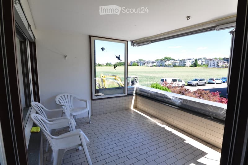 Balcony with tiled floor, white plastic chairs, and a view of a field and buildings.