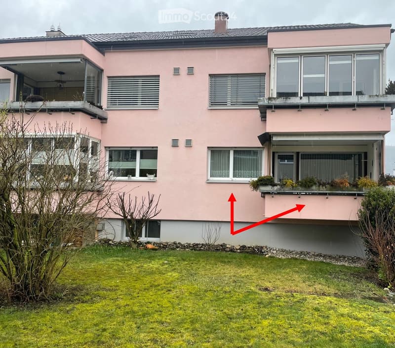 Two-story pink house with multiple windows, balconies, and a raised ground floor. The house has a well-maintained lawn in front.