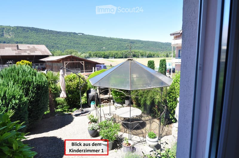A sunny outdoor patio with a round table, chairs, an umbrella, and potted plants, surrounded by greenery and overlooking a scenic countryside view.