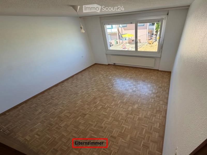 Empty room with wooden parquet flooring, white walls, and a large window with a view of the street.