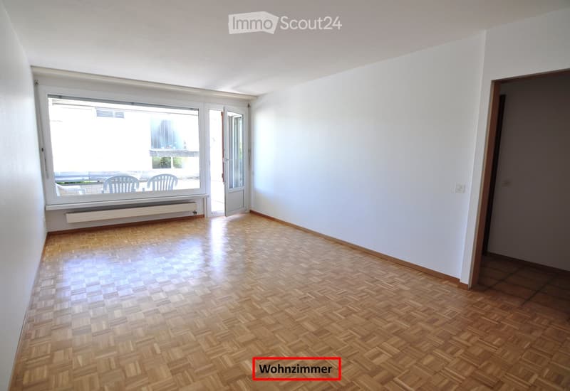 Empty room with wooden parquet flooring, white walls, large window with a view to an outdoor area, and a door leading to the balcony.