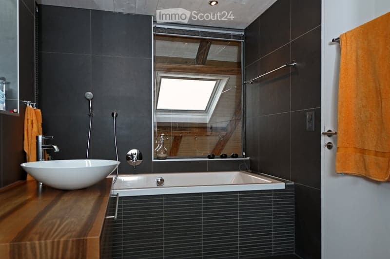 Modern bathroom with dark grey tiles, a white sink, a bathtub, a large mirror, and an orange towel hanging on the wall.