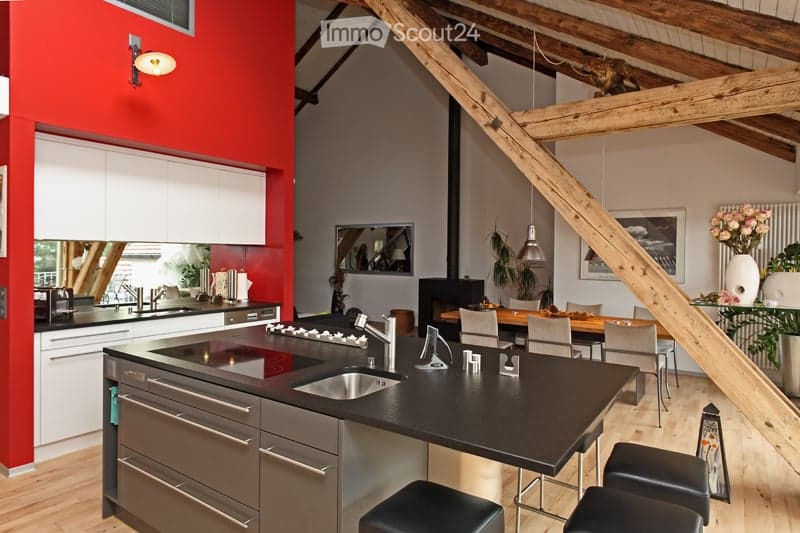 Modern kitchen with a black countertop island, stainless steel sink, faucet, stove, and cabinets. There is a dining area with a wooden table, chairs, and a flower vase on the counter. The space features exposed wooden beams and pendant lighting.