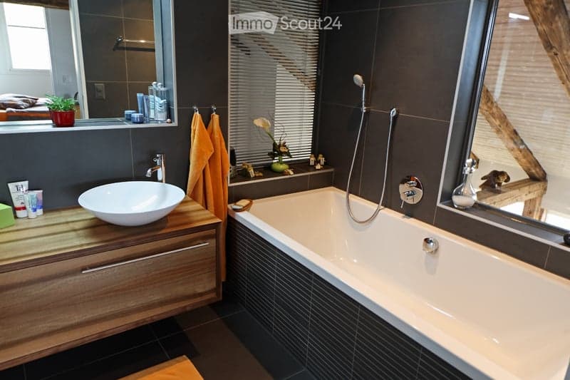 Modern bathroom with a large bathtub, a wooden vanity with a white sink, a mirror, and a towel hanging on a rack. The room features dark tiles and a sleek design.