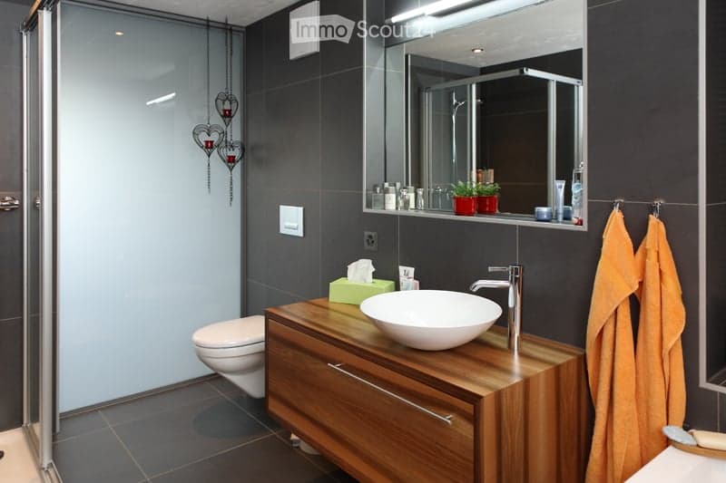 Modern bathroom with dark tiled walls, a wooden vanity with a white sink, a toilet, a large mirror, and decorative hanging hearts with candles. Orange towels are neatly hung on a rack.
