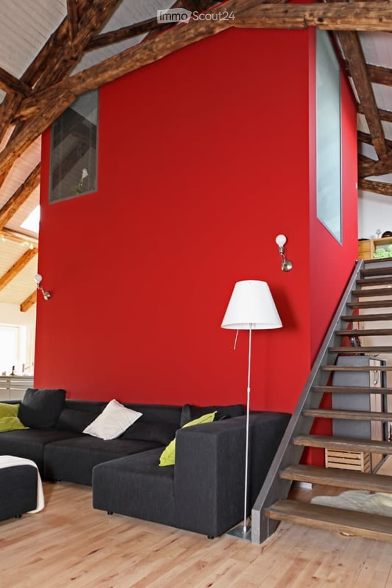 Modern living room with exposed wooden beams, red accent wall, black sectional sofa with green and white pillows, floor lamp, and wooden staircase leading to an upper level.