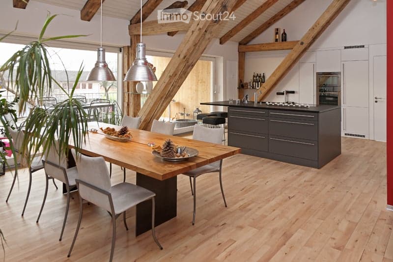 Open-plan kitchen and dining area with light wooden flooring, exposed wooden beams, modern kitchen island with cabinets, dining table with chairs, pendant lights, and decorative bowl on the table.