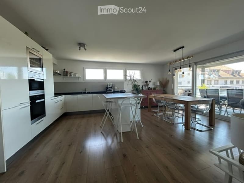 Open-plan kitchen and dining area with light wood flooring, white cabinets, built-in oven, and a dining table with chairs. Large windows offer a view of the city.