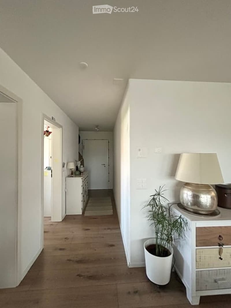 A narrow hallway with wooden flooring, white walls, and a lamp on a small table. There is a potted plant and a small cabinet visible.