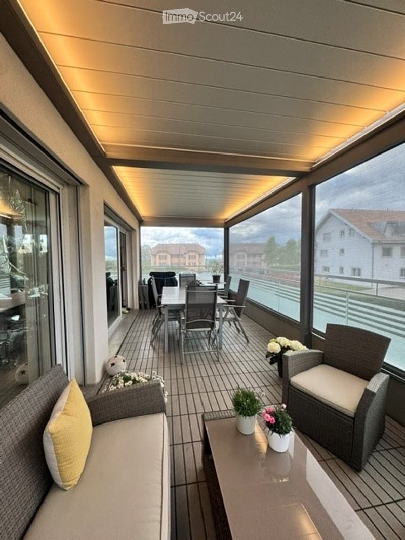 Modern balcony with large windows, tiled floor, and outdoor furniture including a sofa, chairs, and a dining set.