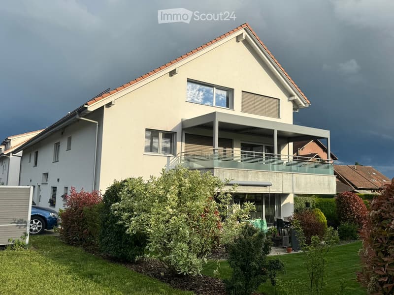 Two-story house with a red-tiled roof, large windows, and a modern balcony with glass railings. The front yard is landscaped with shrubs and a small patio area with outdoor furniture.