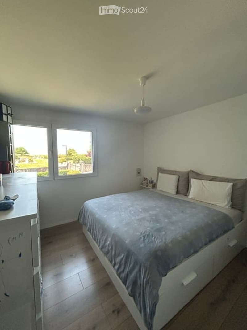 Bright bedroom with wooden flooring, large windows, a bed with grey bedding, nightstand, and a ceiling fan.