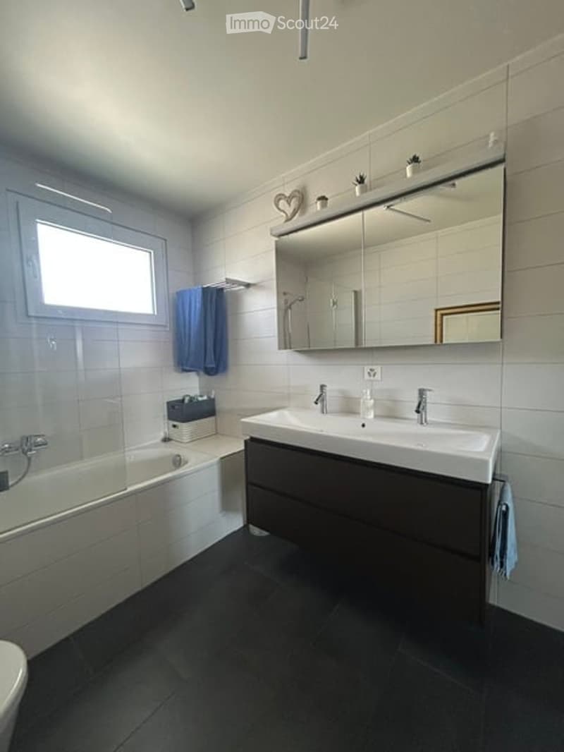 Modern bathroom with white tiles, a bathtub, a double sink with a large mirror, and a shower area. Blue towels are neatly hung on the wall.